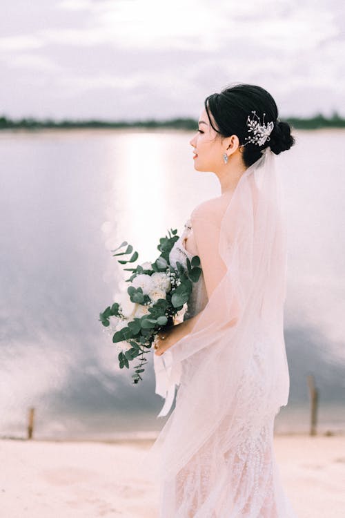 Side view of elegant Asian female in white wedding dress standing with bouquet of flowers in hands on waterfront near river
