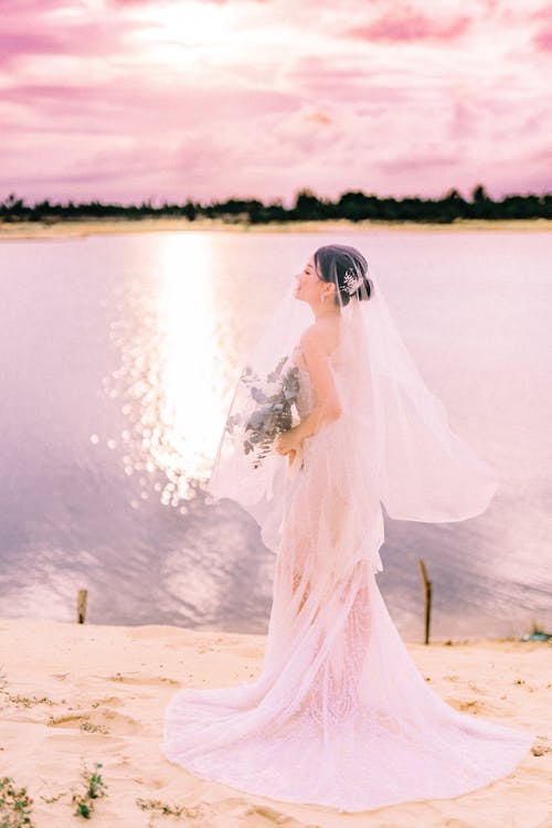 Full body side view of bride in white wedding dress and veil standing on coast with bouquet of flowers in hands near river