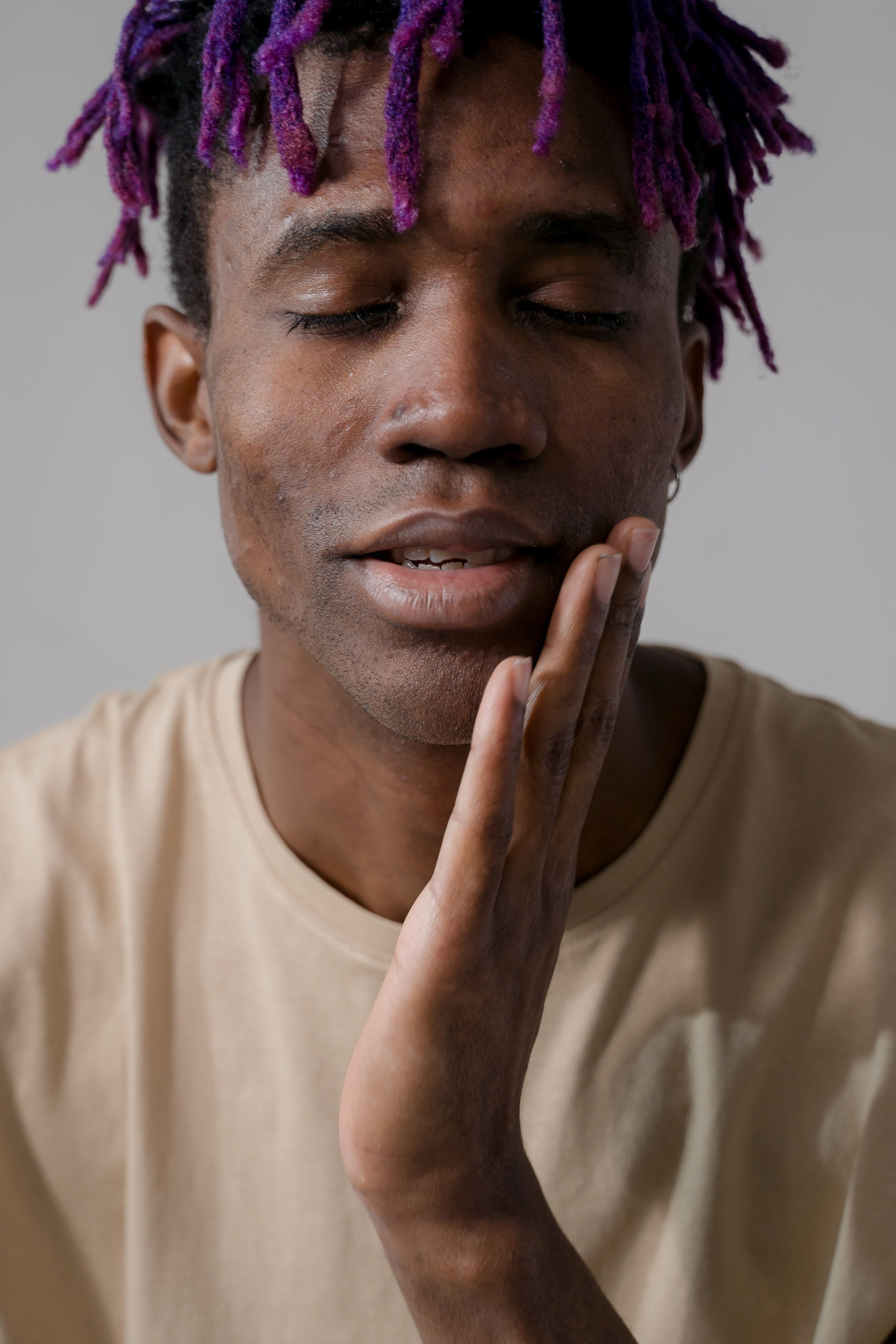 Close-up portrait of an African American man with purple hair and hand on face, eyes closed.