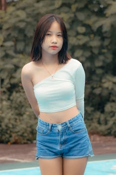 Portrait of a young woman in denim shorts and a crop top, posing outdoors with a lush green background.