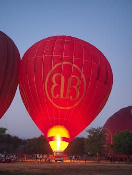 A vibrant red hot air balloon being prepared for takeoff at sunrise with people gathered around in Myanmar.