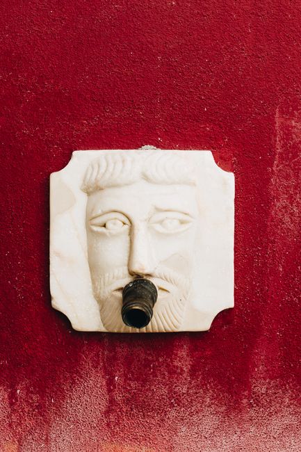 Square marble fountain sculpture depicting a face against a vibrant red wall.