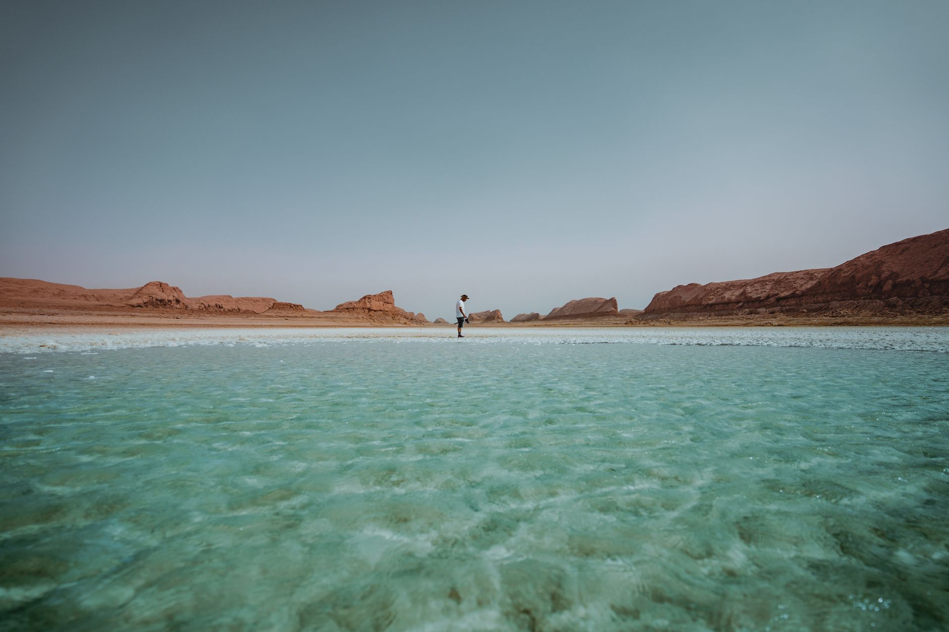 A lone individual stands in a serene turquoise lagoon in Iran's Dasht-e Lut Desert, surrounded by arid landscapes.