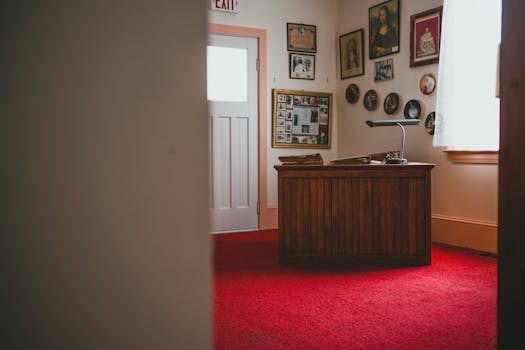 Vintage office interior with red carpet, wooden desk, framed photos, and natural light through window.