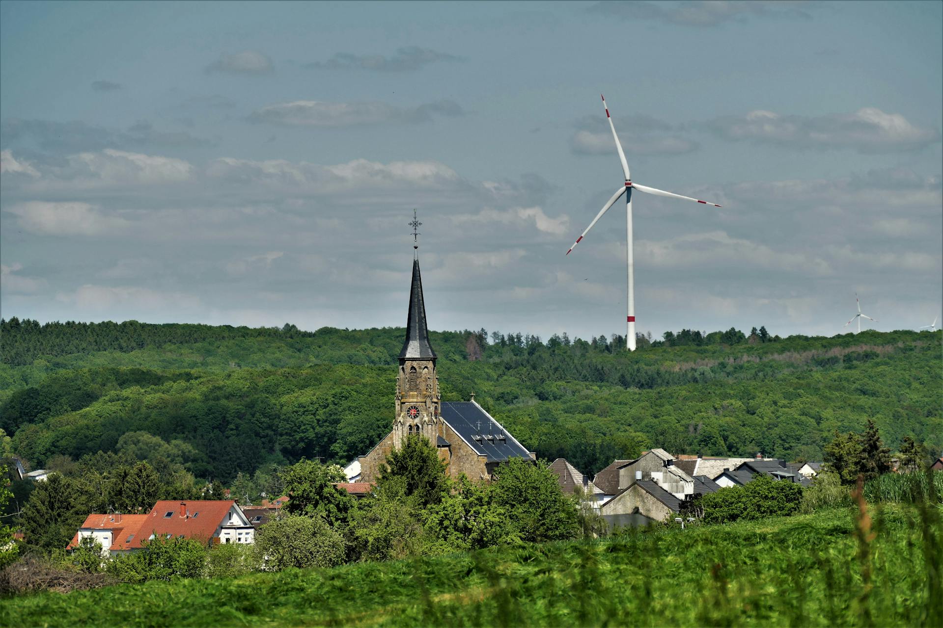 A scenic view of a wind turbine beside a church steeple in the lush, green countryside of Saarwellingen, Germany.