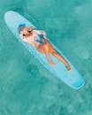 穿着泳衣的金发女人漂浮在蓝色的冲浪板上，在清澈的绿松石水中，闭上眼睛，享受夏日的阳光