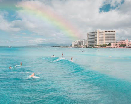 Surfers ride gentle waves beneath a vibrant rainbow at Waikiki Beach with hotels in the background.
