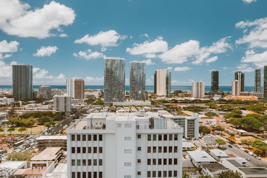 Photo by Jess Loiterton Aerial view of a modern city skyline with the ocean in the background under a blue sky.