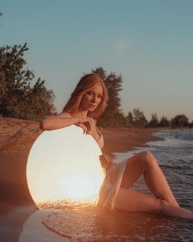 A woman sits on the beach with a mirror, reflecting a warm sunset over the water.