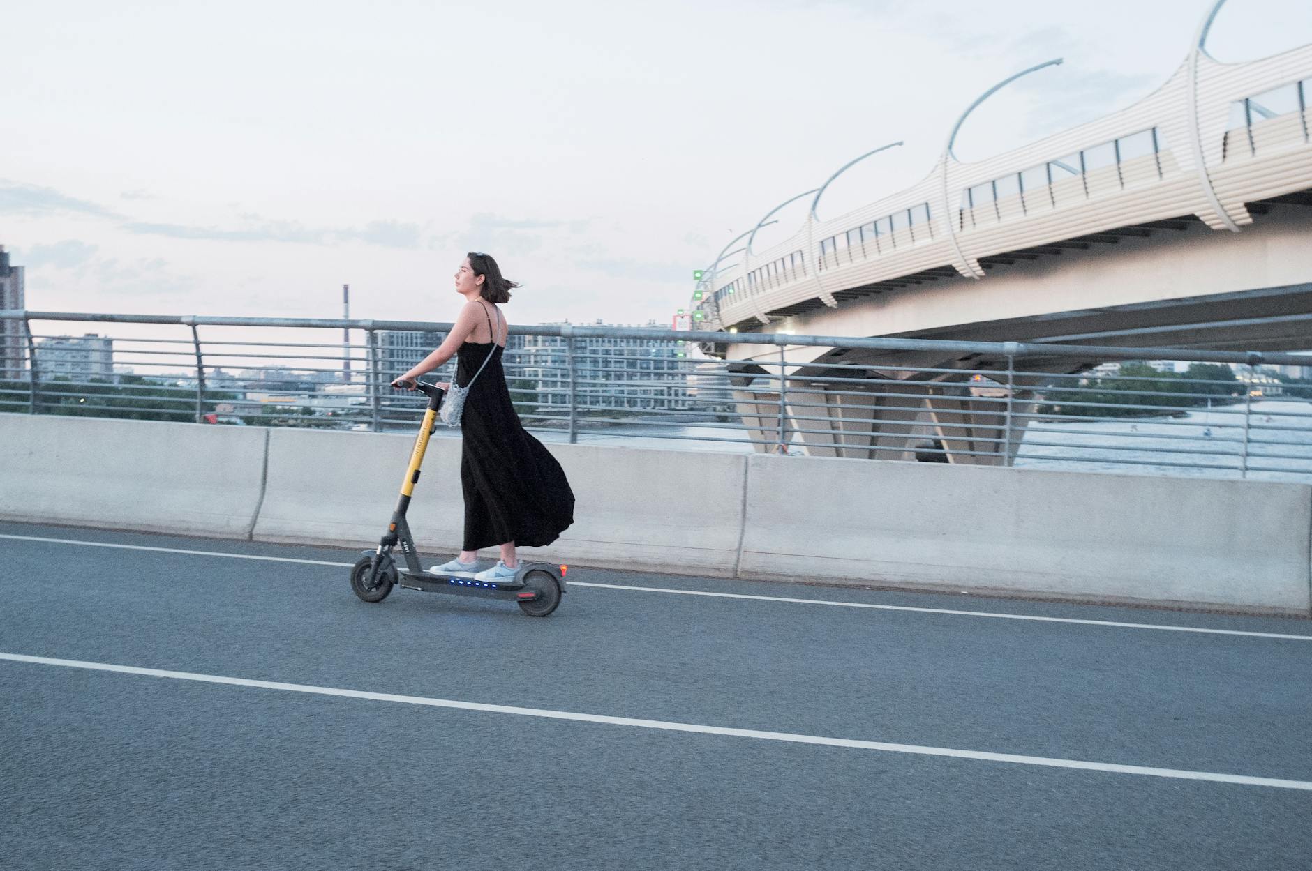 A woman enjoys a ride on an electric scooter across a modern urban bridge at sunset.