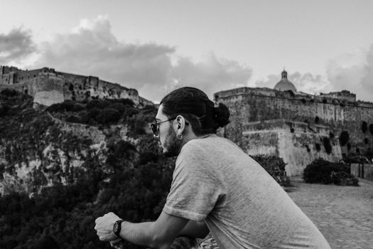 A man leans on a railing observing ancient stone architecture at dusk in black and white.
