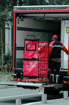 A delivery man is unloading a truck filled with supplies on an urban street, wearing a red and black uniform.