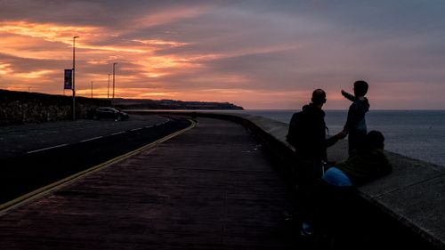Free Silhouetted family watching a sunset over the coastline with a road nearby. Stock Photo