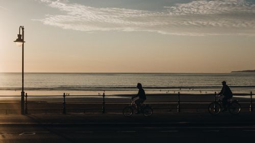 Free Two cyclists ride along a seaside path at dawn, silhouetted against the tranquil ocean horizon. Stock Photo