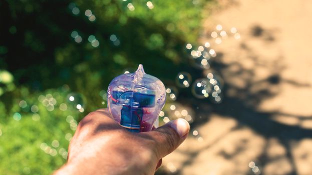 Hand holding a colorful bubble gun outdoors on a sunny day, creating joyful bubbles.