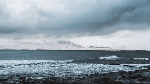 Free Serene winter coastal view with snow-covered mountains and a dramatic cloudy sky. Stock Photo