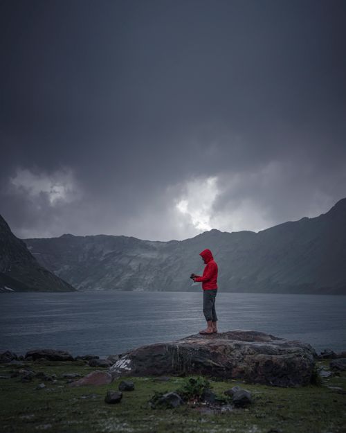 Free A person in a red jacket stands alone on a rock by a large lake under dark storm clouds. Stock Photo