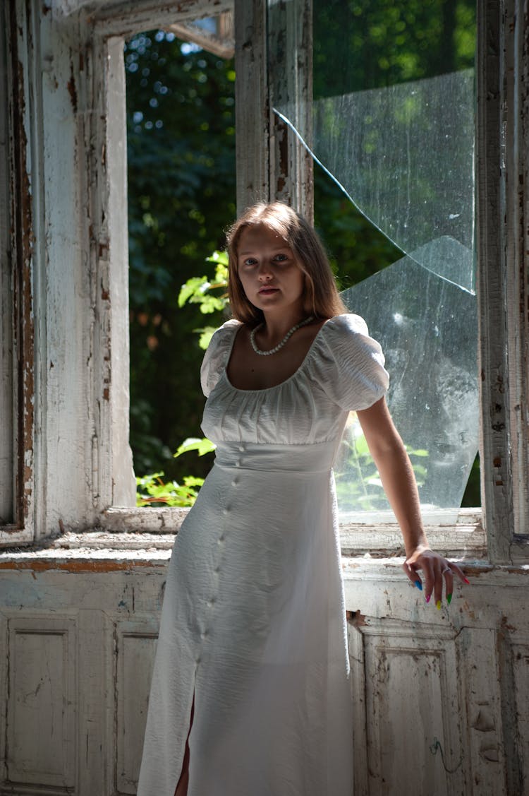 Young Woman In White Dress Standing Near Window With Broken Glass