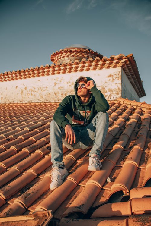 Free A young man sitting on a Mediterranean-style rooftop wearing a green hoodie and sunglasses. Stock Photo