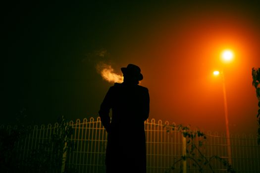 A silhouetted person in a hat smoking under an orange streetlight in a foggy night.