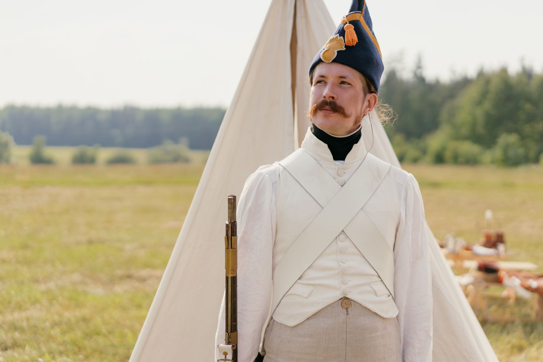 A historical reenactor in Napoleonic military uniform standing by a tent in open countryside.