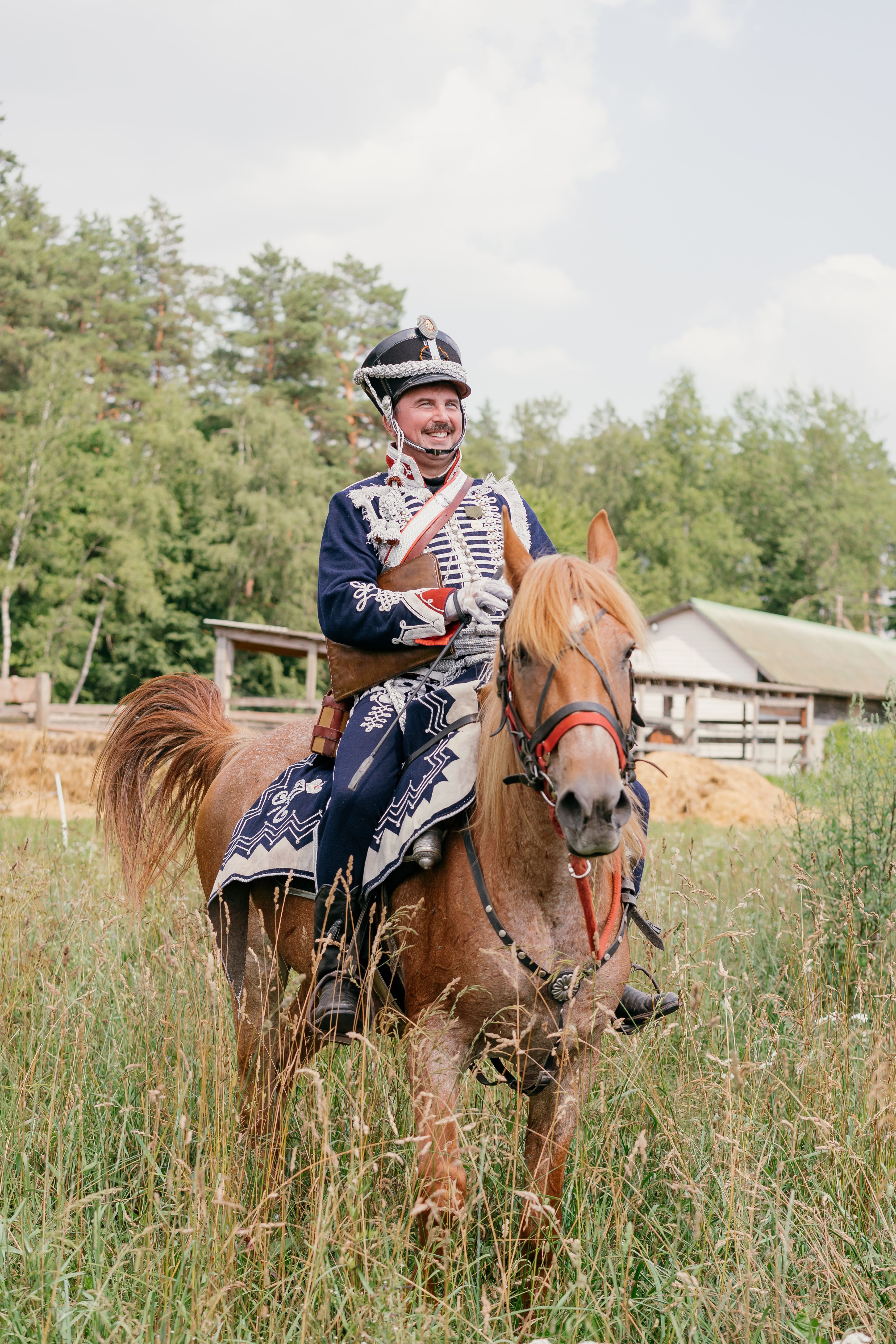 Man in traditional uniform riding a brown horse outdoors, evoking historical charm.