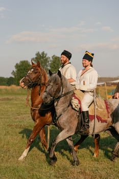 Two soldiers in historical uniforms riding horses in a grass field during reenactment.