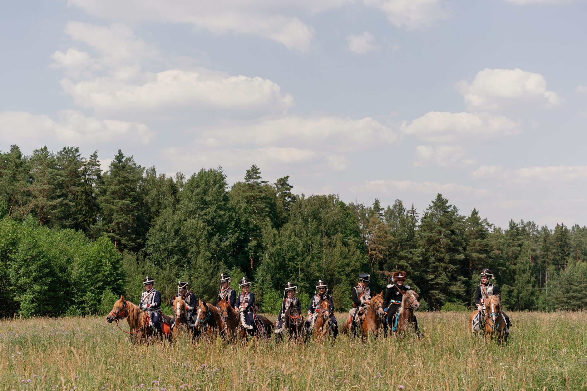 A group of reenactors in historical military uniforms riding horses through a lush countryside field.