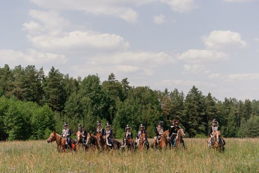 A group of reenactors in historical military uniforms riding horses through a lush countryside field.