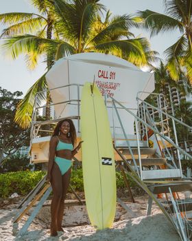 A woman in swimwear poses with a surfboard next to a lifeguard tower on a tropical beach.