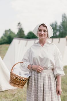 Woman in historical attire holding a basket outdoors, with tents in the background.