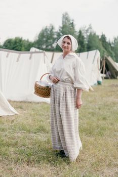 Woman in historical attire standing in a meadow, holding a wicker basket by tents.