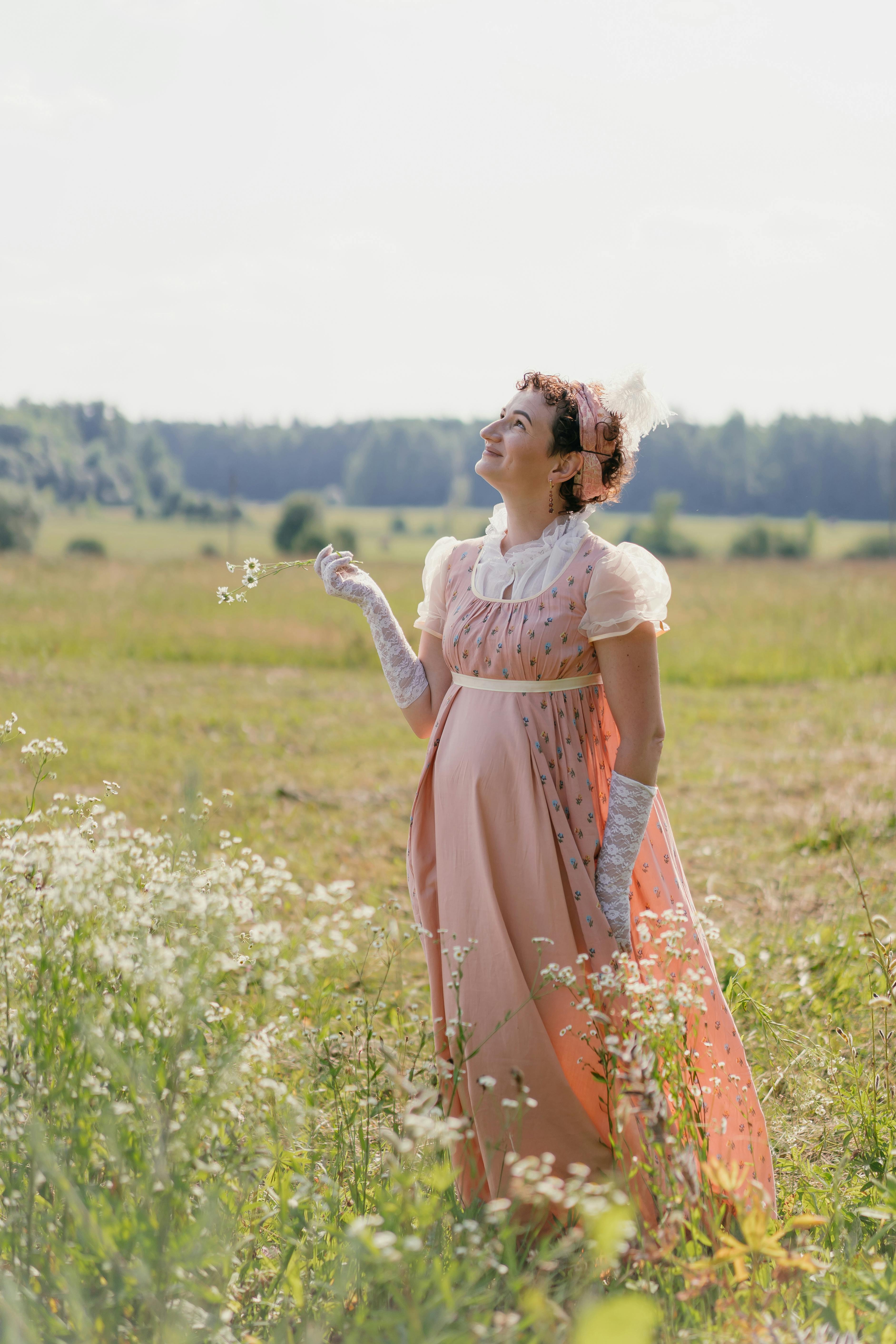 Woman in a vintage gown holding flowers in a sunny meadow, smiling upwards.