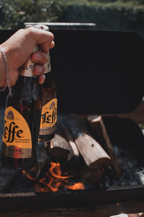 Closeup of a Hand Holding Beer Bottles and Grill with Log on Fire in Background