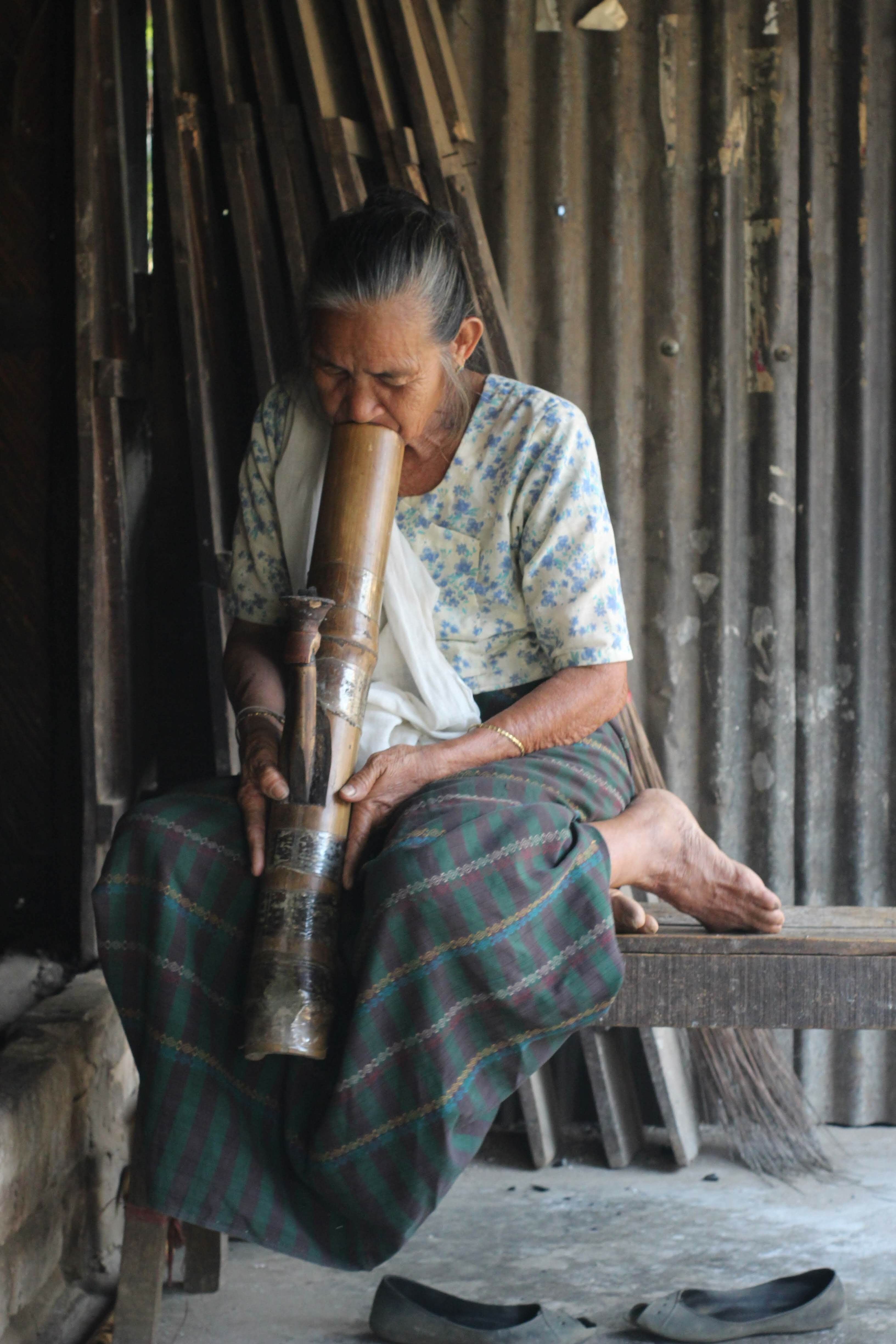 Elderly woman smoking from a bamboo pipe in a traditional setting indoors.