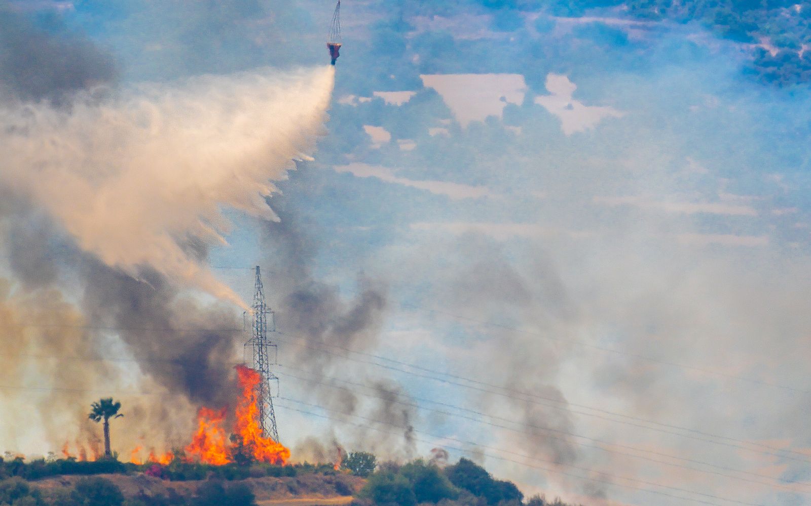 A helicopter carrying a water bucket flying over a forest fire.