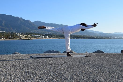 A man performs a yoga pose on a seafront with stunning mountain views under a clear blue sky.