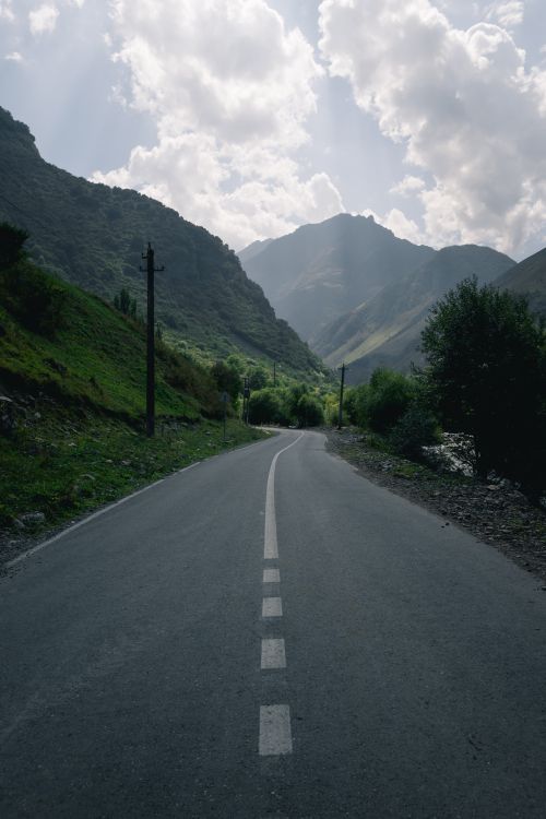 Free A peaceful empty road winding through lush green mountains under a cloudy sky. Stock Photo