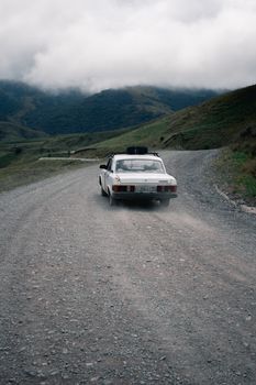 A white car navigates an unpaved road in a mountainous area under cloudy skies.