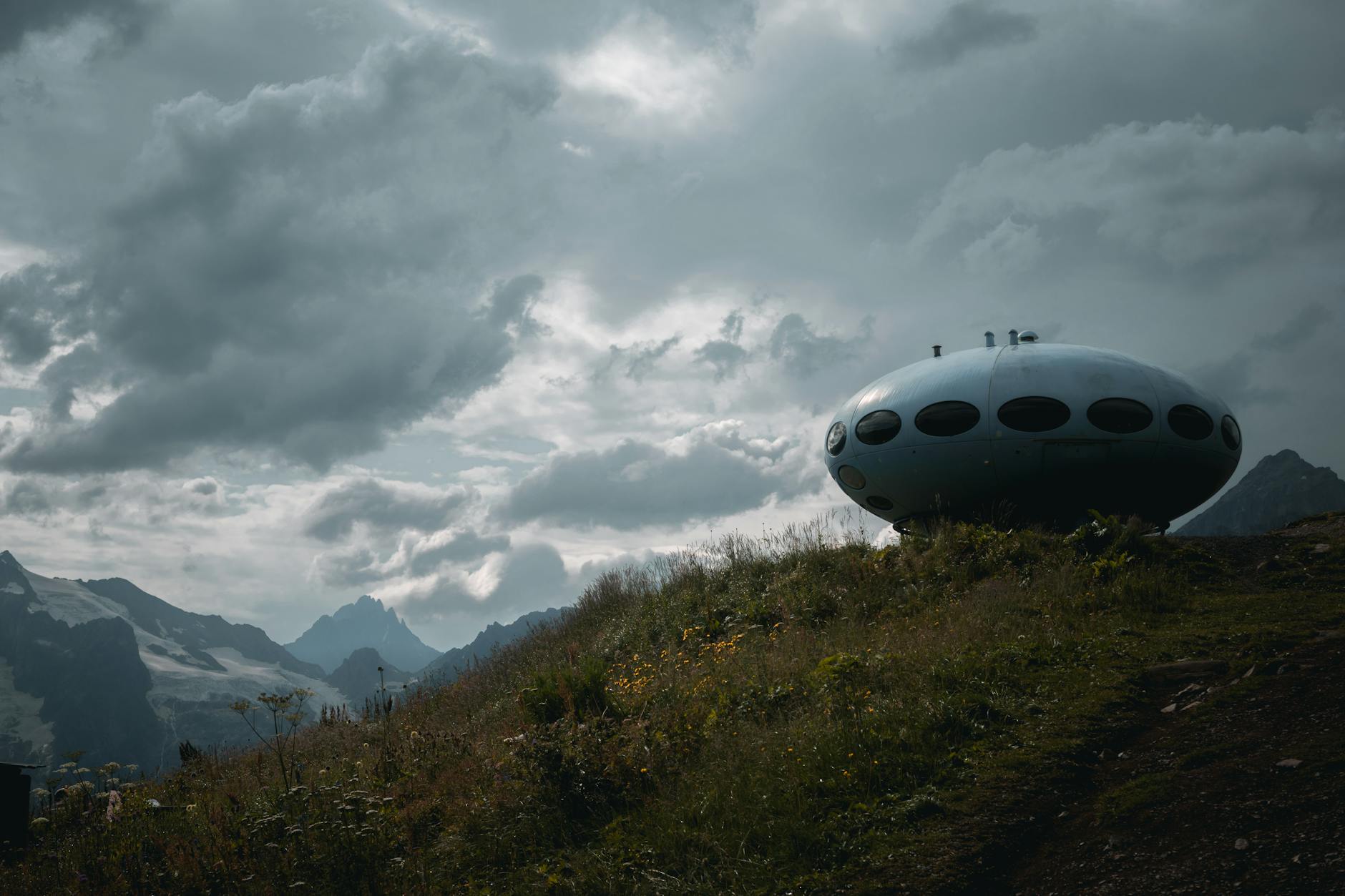 Unique UFO-like structure on a grassy hill with a cloudy mountain backdrop.