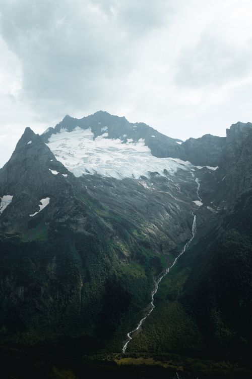 Free A stunning view of a snow-capped mountain with a flowing stream below, captured under an overcast sky. Stock Photo