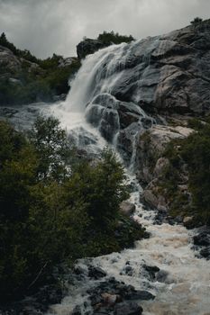 A scenic waterfall flowing over rocky terrain amidst lush greenery, captured on a cloudy day.