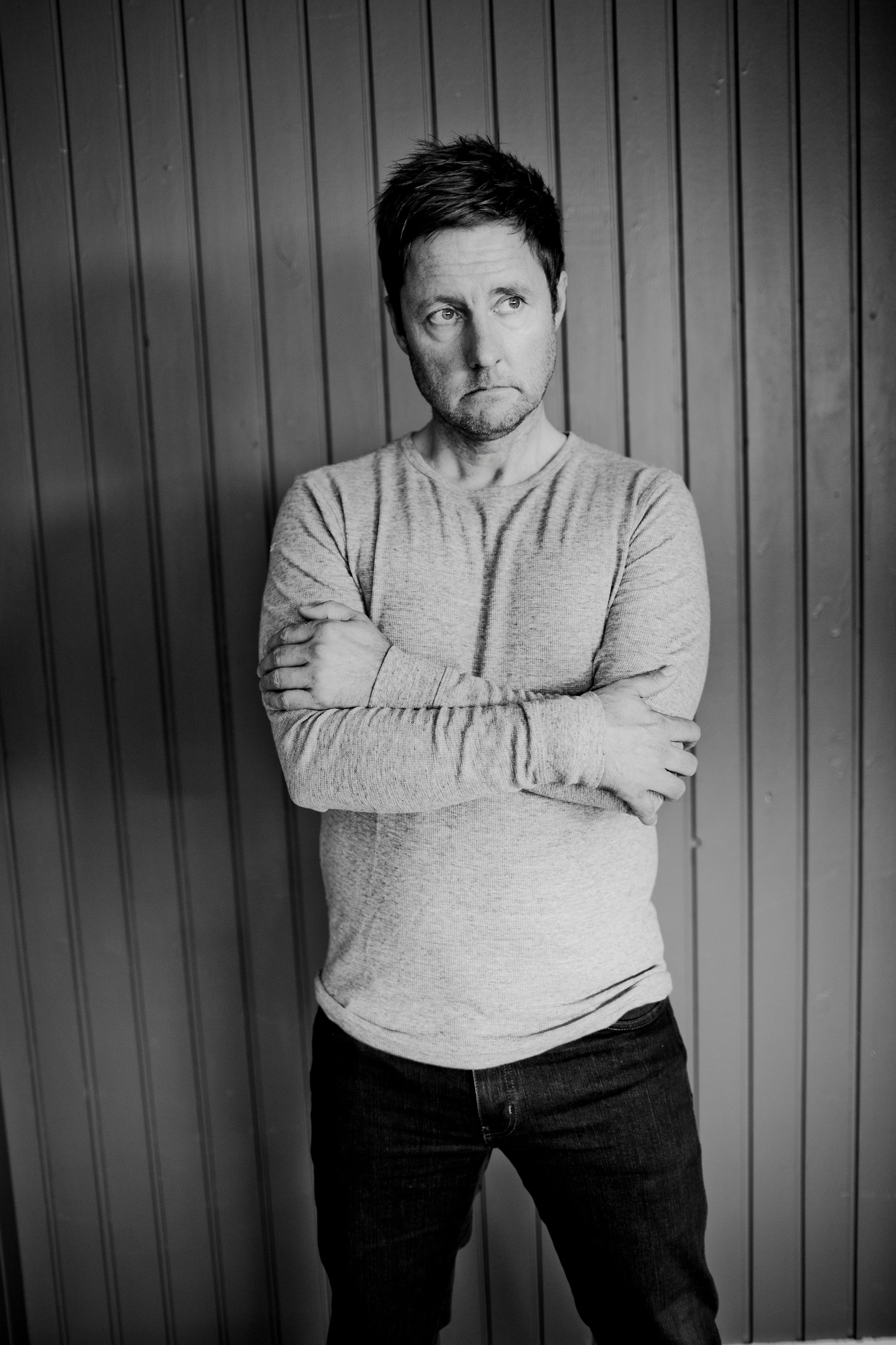 A man in a long sleeve shirt stands pensively against a wooden backdrop.