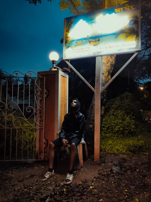 Full body of young Indian male traveler in hooded jacket relaxing on chair near metal fence and street banner in park in evening