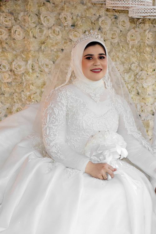 Free A beautiful bride in a white traditional gown, sitting elegantly before flower backdrop. Stock Photo