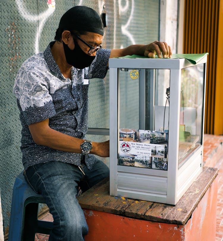 Man Wearing A Face Mask Sitting On A Chair On A Sidewalk With A Glass Container 