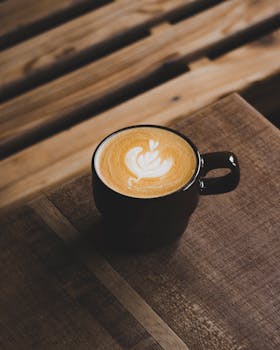 A stylish cappuccino with intricate latte art in a black cup on a rustic wooden table.