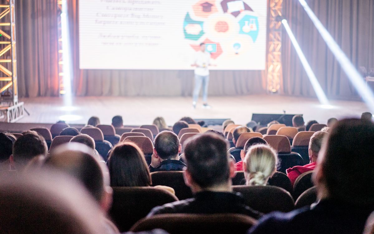Audience attentively watching a presentation in a conference setting, with stage lights and speaker visible.