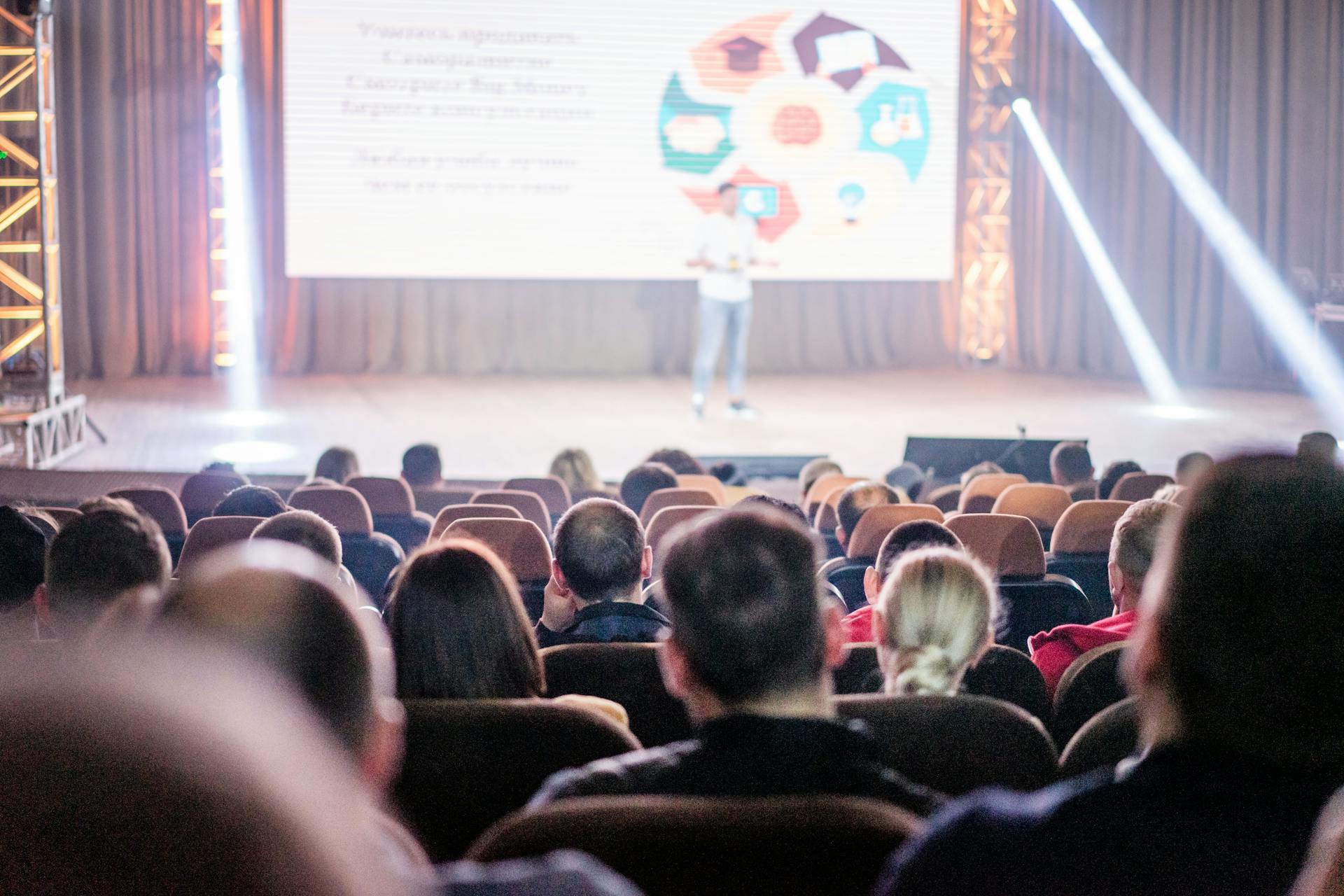 Audience attentively watching a presentation in a conference setting, with stage lights and speaker visible.