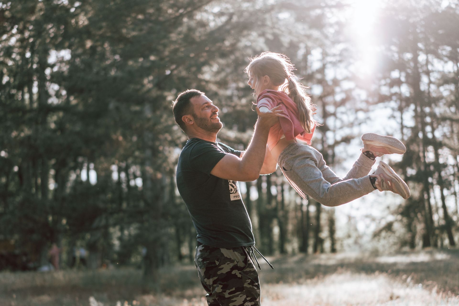 A father lifting his laughing daughter into the air outdoors with trees in the background